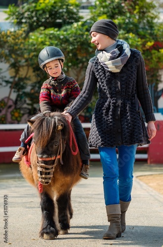 Photo: Mother riding her son on a pony wearing protective helmet Mother riding her son on a pony wearing protective helmet