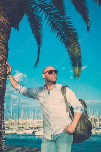 Photo: Middle-aged man with backpack standing near palm tree against ya Middle-aged man with backpack standing near palm tree against ya
