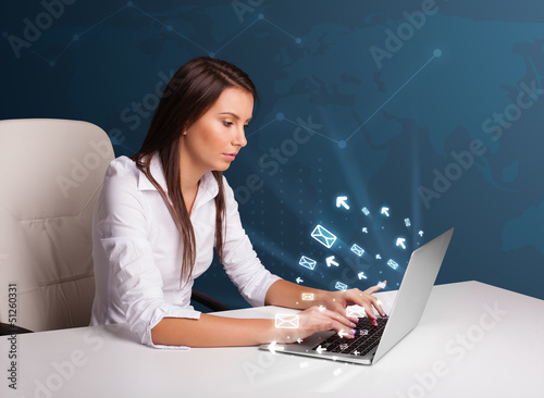 Photo: Young lady sitting at dest and typing on laptop with message ico Young lady sitting at dest and typing on laptop with message ico