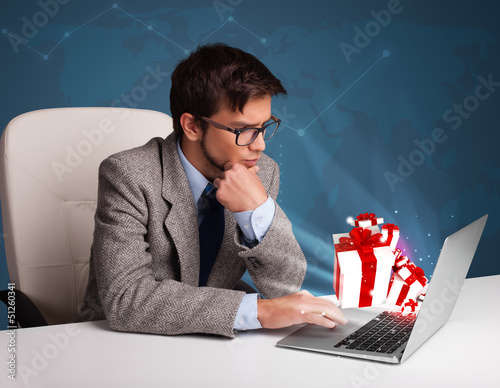 Photo: Handsome man sitting at desk and typing on laptop with present b Handsome man sitting at desk and typing on laptop with present b