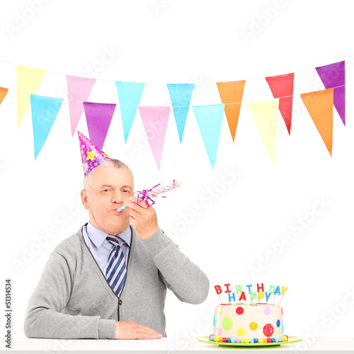 Photo: A happy mature man with party hat blowing and a birthday cake A happy mature man with party hat blowing and a birthday cake