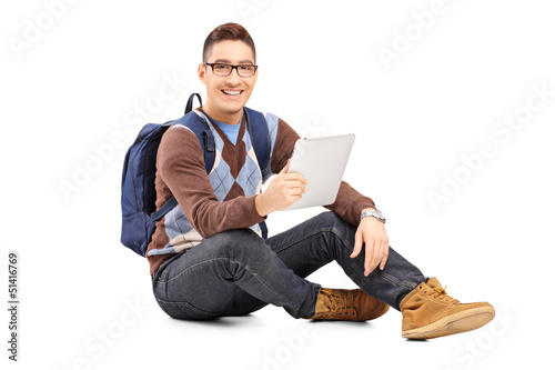 Photo: Smiling male student with backpack seated on a floor holding a t Smiling male student with backpack seated on a floor holding a t