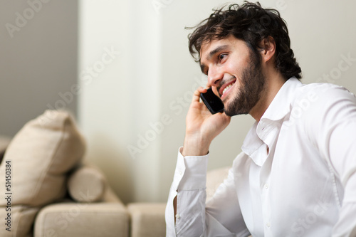Photo: Young man smiling and talking on smart phone at home Young man smiling and talking on smart phone at home