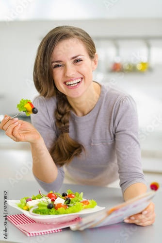 Photo: Smiling young housewife eating fresh salad and reading magazine Smiling young housewife eating fresh salad and reading magazine