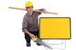 Man kneeling by sign holding saw and plank of wood