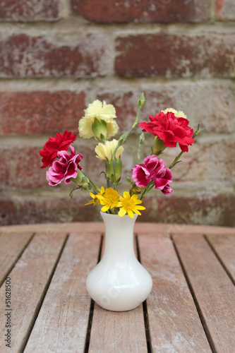 Photo: Carnations and celandines in white vase on garden table Carnations and celandines in white vase on garden table