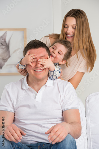 Photo: Happy family having fun in the living room: son surprisng his fa Happy family having fun in the living room: son surprisng his fa