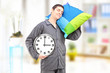 Young lazy guy holding a clock and sleeping on a pillow at home