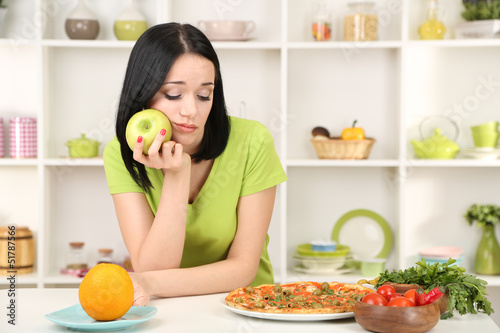Photo: Pretty girl selects pizza or diet on kitchen background Pretty girl selects pizza or diet on kitchen background