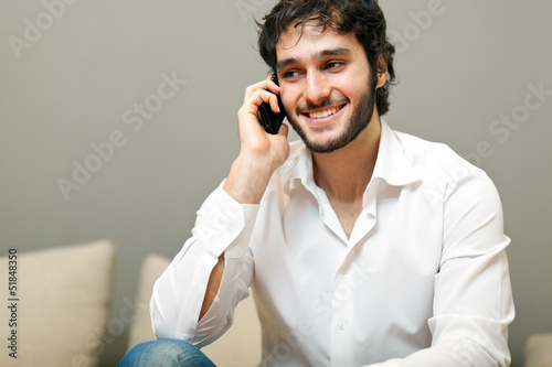 Photo: Young man smiling and talking on smart phone at home Young man smiling and talking on smart phone at home