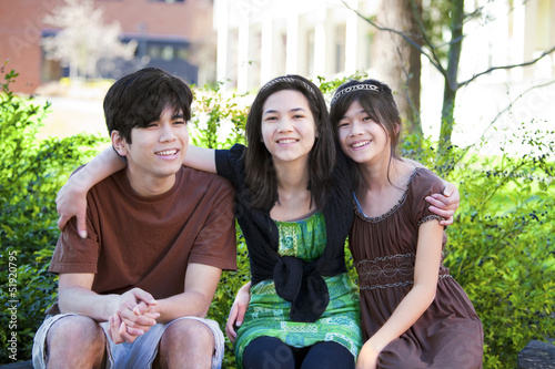 Photo: Three brother and sisters sitting outdoors on log, smiling Three brother and sisters sitting outdoors on log, smiling