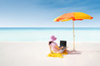 Asian woman typing on laptop under parasol at beach