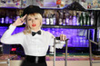 Glamorous blonde girl in black peaked cap sits near bar counter.