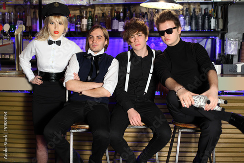 Photo: Four young rock band in black and white pose near bar counter. Four young rock band in black and white pose near bar counter.