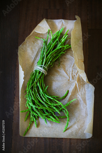 Photo: Green wild asparagus bunch on paper and brown wooden desk Green wild asparagus bunch on paper and brown wooden desk