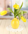 Yellow tulips in vase on wooden table on room background
