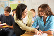 Teacher Helping Female Pupil Studying At Desk In Classroom