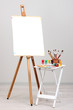 Wooden easel with clean paper and art supplies in room