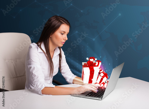 Photo: Pretty lady sitting at desk and typing on laptop with present bo Pretty lady sitting at desk and typing on laptop with present bo