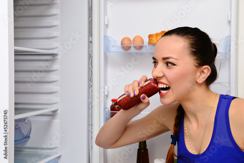 Photo: Girl eats sausage near the refrigerator Girl eats sausage near the refrigerator