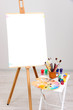 Wooden easel with clean paper and art supplies in room