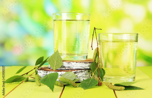 Photo: Glasses of birch sap on wooden table, on green background Glasses of birch sap on wooden table, on green background