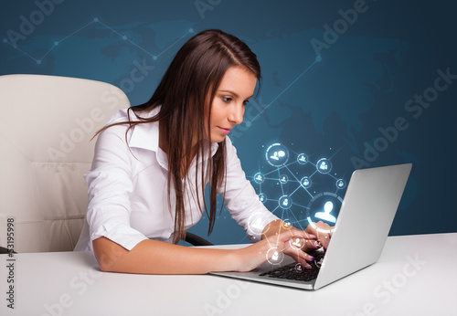 Photo: Young lady sitting at desk and typing on laptop with social netw Young lady sitting at desk and typing on laptop with social netw