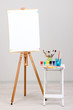 Wooden easel with clean paper and art supplies in room