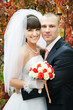 Groom hugging bride against blurred autumn red leaves