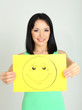 Young woman holding paper with happy smiley on gray background.