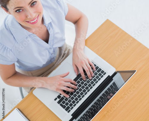 Photo: Pretty businesswoman typing on her laptop and smiling up at came Pretty businesswoman typing on her laptop and smiling up at came
