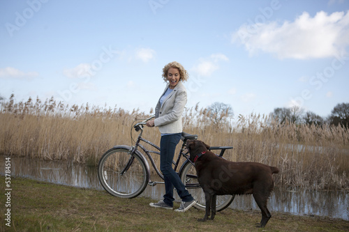 Photo: A mid adult woman pushing a bicycle next to a lake with her dog A mid adult woman pushing a bicycle next to a lake with her dog