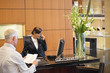 Photo: Businessman reading a brochure with a receptionist talking on a telephone at the hotel reception counter Businessman reading a brochure with a receptionist talking on a telephone at the hotel reception counter