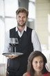 Photo: Woman sitting in a restaurant with a waiter holding a tray of wine glasses in the background Woman sitting in a restaurant with a waiter holding a tray of wine glasses in the background