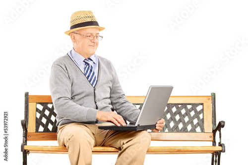 Photo: Senior man sitting on a wooden bench and working on a laptop Senior man sitting on a wooden bench and working on a laptop