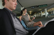Photo: Businesswoman text messaging on phone in airport lounge, businessman reading newspaper Businesswoman text messaging on phone in airport lounge, businessman reading newspaper