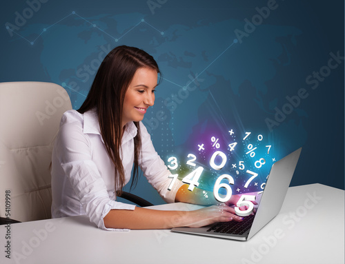 Photo: Beautiful young woman sitting at desk and typing on laptop with Beautiful young woman sitting at desk and typing on laptop with