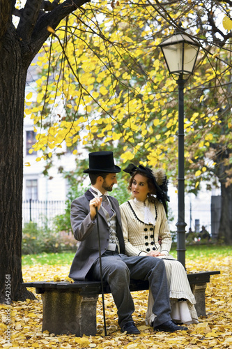 Photo: Old-fashioned dressed couple on a park bench in fall. Old-fashioned dressed couple on a park bench in fall.