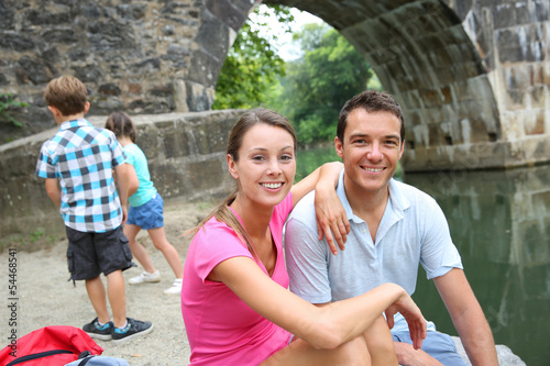 Photo: Couple relaxing by bridge, kids in background Couple relaxing by bridge, kids in background