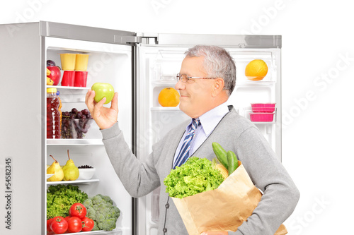 Photo: Smiling mature man holding a paper bag near the refrigerator ful Smiling mature man holding a paper bag near the refrigerator ful
