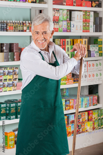 Photo: Senior Male Owner Standing Against Shelves In Supermarket Senior Male Owner Standing Against Shelves In Supermarket