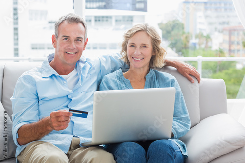 Photo: Happy couple sitting on their couch using the laptop to buy onli Happy couple sitting on their couch using the laptop to buy onli