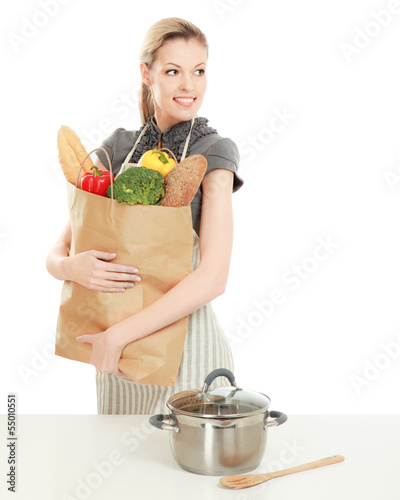 Photo: Woman in apron holding grocery bag, standing near table with pan Woman in apron holding grocery bag, standing near table with pan