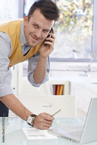 Photo: Young man talking on mobile in office, smiling Young man talking on mobile in office, smiling