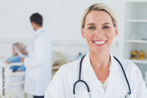 Photo: Smiling blond doctor posing with doctor attending patient on bac Smiling blond doctor posing with doctor attending patient on bac