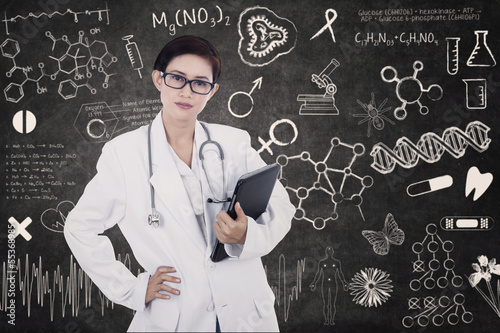 Photo: Confident female doctor hold e-tablet on written blackboard Confident female doctor hold e-tablet on written blackboard