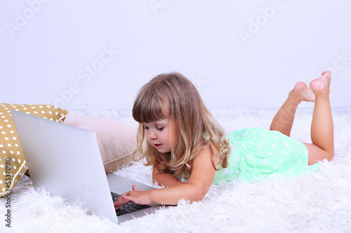 Photo: Little cute girl lies on carpet with laptop, on gray background Little cute girl lies on carpet with laptop, on gray background