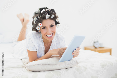 Photo: Smiling brunette in hair rollers lying on her bed using her tabl Smiling brunette in hair rollers lying on her bed using her tabl