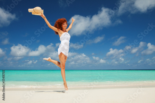 Photo: Happy young woman in white dress with hat jumping on the beach Happy young woman in white dress with hat jumping on the beach