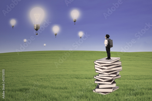 Photo: Business kid stand on books looking at lightbulbs outdoor Business kid stand on books looking at lightbulbs outdoor